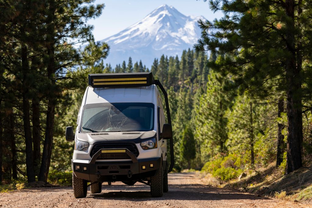 A rugged off-road van is parked on a forest trail surrounded by pine trees, with a snow-capped mountain rising in the background under clear daylight, conveying durability and adventure-ready capability.