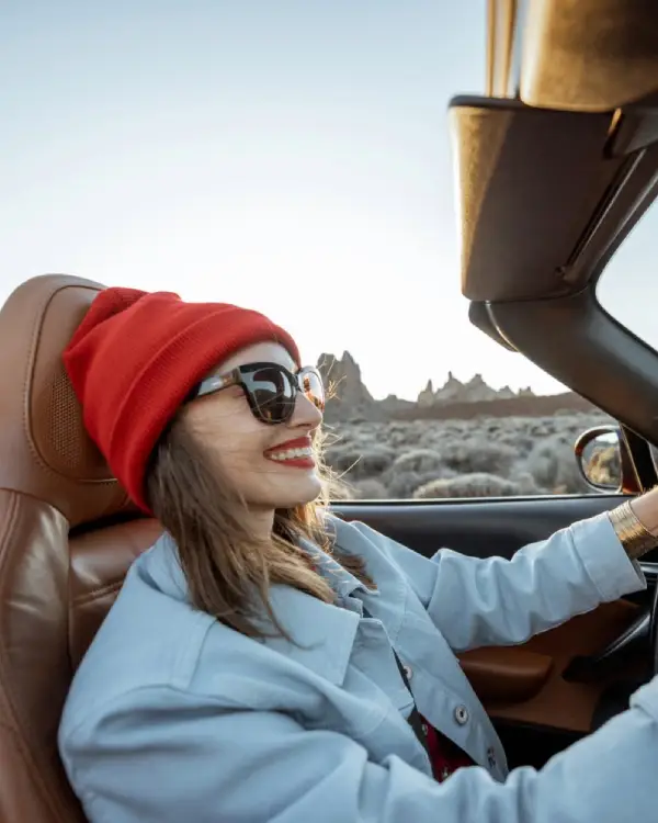 A smiling woman wearing a red beanie and sunglasses drives a convertible through a scenic desert landscape under a clear sky.