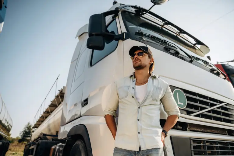 A man wearing sunglasses and a cap stands in front of a large commercial truck, representing professional fleet operations in a transport or logistics setting.