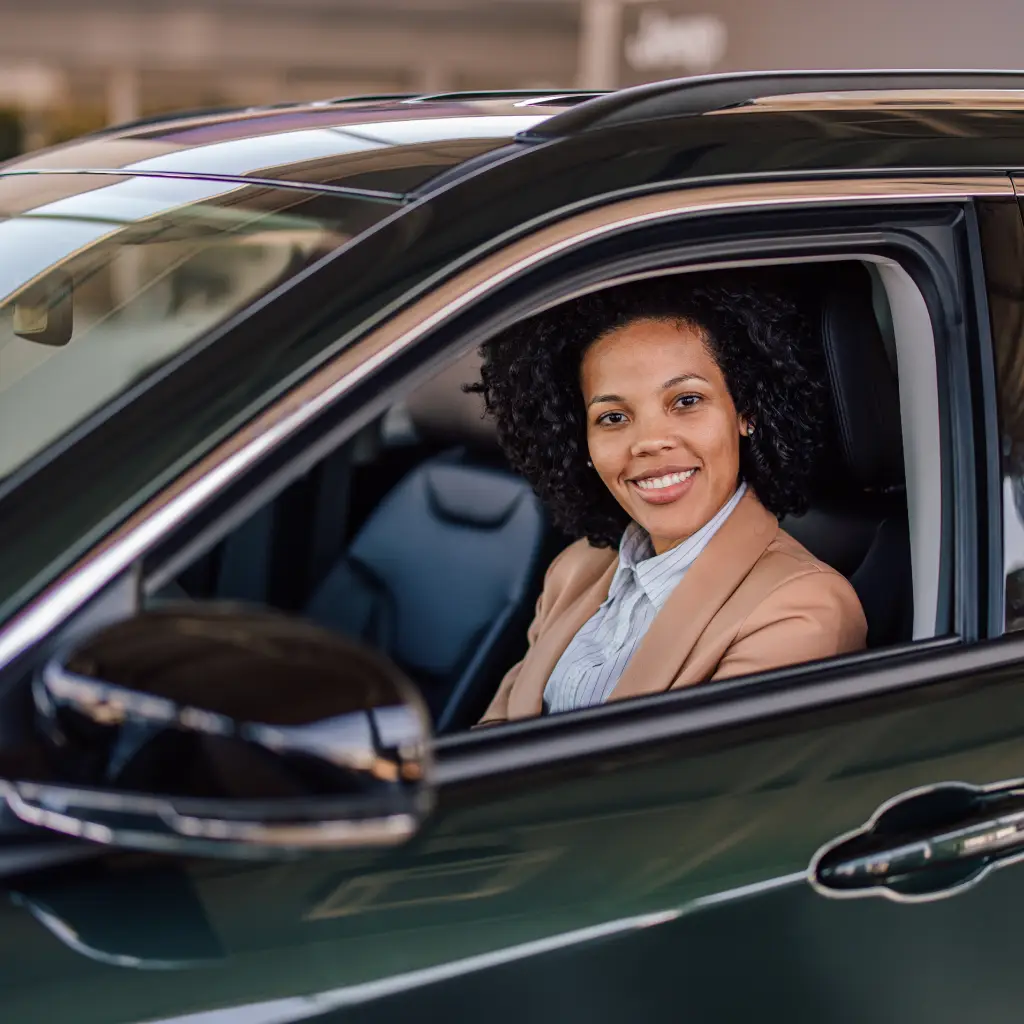Professional woman smiling from the driver’s seat of a dark-colored SUV, wearing a tan blazer and light blouse, with the car window open in a dealership setting.