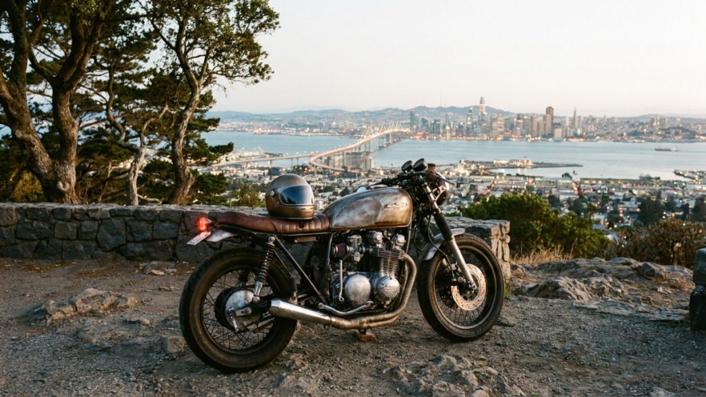 Side view of a raw metal custom cafe racer with a brown leather seat and a helmet resting on the tank, parked on a rocky hilltop with a panoramic view of the San Francisco Bay, the Bay Bridge, and the city skyline at dusk